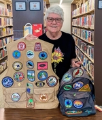 Inside Library, Background: Bookshelves, Jan Lohmann Holding Tan Vest And Backpack With Patches